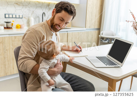 Self-employed young man working from home office while talking care of a baby 127355625