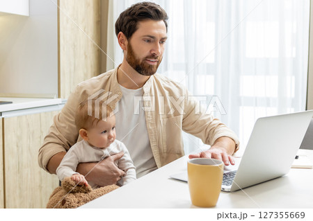 Bearded young man sitting at the laptop and holding his baby boy Bearded young man sitting at the laptop and holding his baby boy 127355669