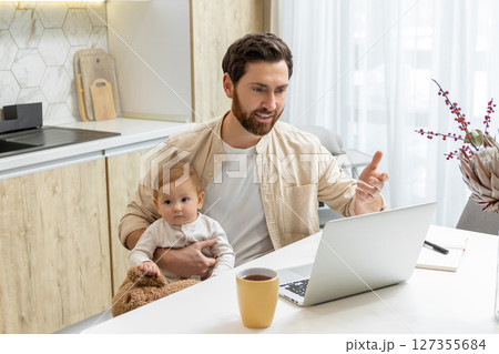 Bearded young man sitting at the laptop and holding his baby boy Bearded young man sitting at the laptop and holding his baby boy 127355684