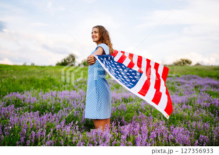 Young woman proudly hold waving american USA flag on blooming meadow. 4th of July. Independence Day. Young woman proudly hold waving american USA flag on blooming meadow. 4th of July. Independence Day. 127356933