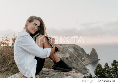 Woman Sunset Cliffside Portrait - A woman in a white shirt sits on a cliff overlooking the ocean at sunset, smiling at the camera. 127358318