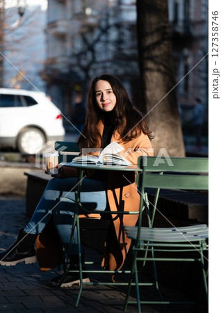Sunlit afternoon at a city cafe with a young woman enjoying a book and coffee in a peaceful outdoor setting surrounded by urban charm 127358746