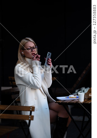 Elegant woman enjoys a moment of focused creativity at a cozy street cafe while sipping coffee and engaging with her phone under soft evening light 127358883
