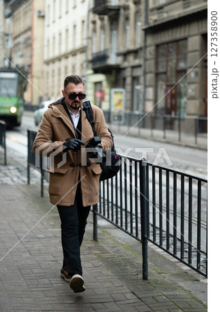 Stylish man walking through a city street, capturing moments with a camera while dressed warmly in winter attire amid a charming urban backdrop Stylish man walking through a city street, capturing moments with a camera while dressed warmly in winter attire amid a charming urban backdrop 127358900