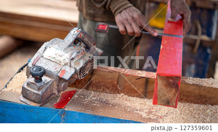 Rustic electric planer on workbench with carpenter hand squaring timber behind 127359601