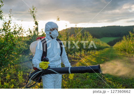 Farmer spraying pesticide in orchard, disinfecting trees in protective suit. Farmer spraying pesticide in orchard, disinfecting trees in protective suit. 127360662