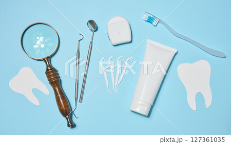 Set of oral hygiene products and tools, tube of toothpaste, dental floss, dental instruments, magnifying glass with heart symbol and paper teeth, blue background. Top view. Set of oral hygiene products and tools, tube of toothpaste, dental floss, dental instruments, magnifying glass with heart symbol and paper teeth, blue background. Top view. 127361035