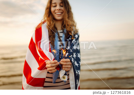 Young woman with American flag and sparklers on beach. Patriotic holiday. USA celebrate 4th of July. 127361816