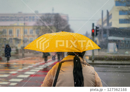 Woman with yellow umbrella standing at city crosswalk in snowy weather, urban winter vibe. Bright yellow umbrella creates vivid contrast in city. Concept of winter commute, solitude, splash of color 127362188