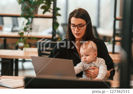 Sitting by laptop. Young mother with her little baby son is in the office 127362531