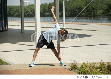 Fit handsome brunette man wearing sports casual clothes and sunglasses doing warm up exercise on city modern outdoor public park. Pause in work. Summer activity. Healthy lifestyle. Fit handsome brunette man wearing sports casual clothes and sunglasses doing warm up exercise on city modern outdoor public park. Pause in work. Summer activity. Healthy lifestyle. 127362925