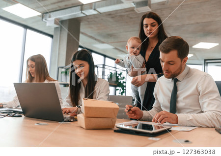 Little boy is in the hands of woman that is in black dress. Infant baby is in the office where group of people are working together 127363038