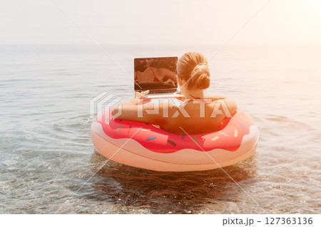 Woman Laptop Beach - Working Remotely on Vacation, Sitting in Inflatable Ring on Water, Sun Setting in the Background. 127363136