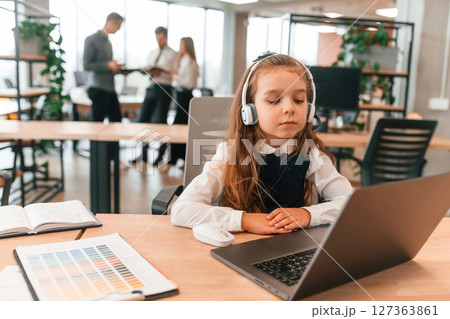 Sitting by the table with laptop. Little girl is in the office where people working together 127363861