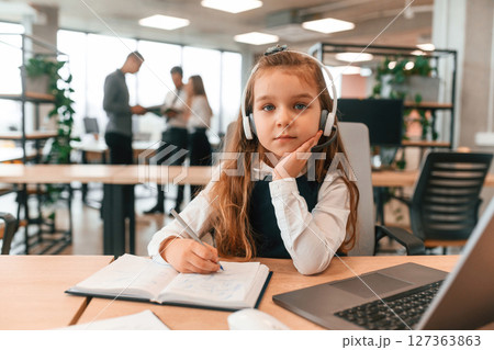 Sitting by the table with laptop. Little girl is in the office where people working together Sitting by the table with laptop. Little girl is in the office where people working together 127363863