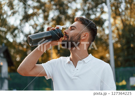 Drinking water, taking a break. Young man is on the tennis court at sunny daytime 127364225