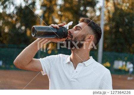 Drinking water, taking a break. Young man is on the tennis court at sunny daytime 127364226