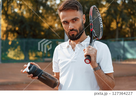 Holding bottle with water and racket. Young man is on the tennis court at sunny daytime Holding bottle with water and racket. Young man is on the tennis court at sunny daytime 127364227