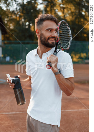Holding bottle with water and racket. Young man is on the tennis court at sunny daytime Holding bottle with water and racket. Young man is on the tennis court at sunny daytime 127364229