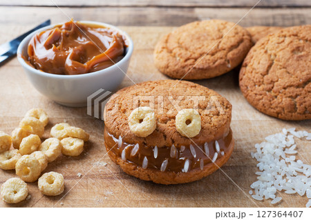 Cookies with cream paste in the shape of monsters for Halloween celebration. Funny homemade faces made of oatmeal cookies and boiled condensed milk. Cookies with cream paste in the shape of monsters for Halloween celebration. Funny homemade faces made of oatmeal cookies and boiled condensed milk. 127364407