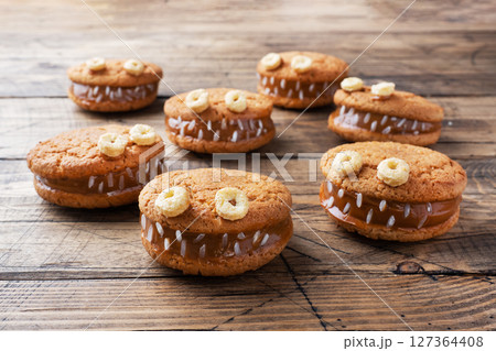 Cookies with cream paste in the shape of monsters for Halloween celebration. Funny homemade faces made of oatmeal cookies and boiled condensed milk. 127364408