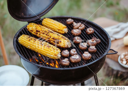 Three ears of corn and several mushrooms grilling on a compact barbecue outdoors, creating a delicious summer meal Three ears of corn and several mushrooms grilling on a compact barbecue outdoors, creating a delicious summer meal 127367869