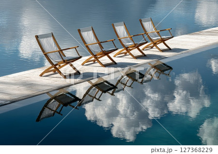 Wooden lounge chairs lined up by tranquil pool reflecting clouds and blue sky 127368229