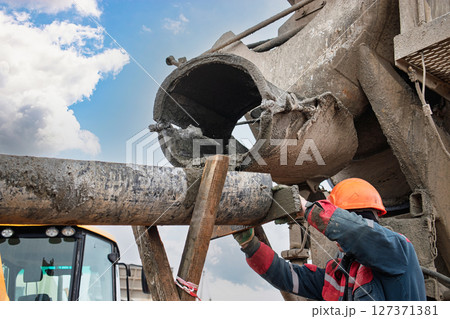 Worker pours concrete from mixer into construction pipe at job site under cloudy sky Worker pours concrete from mixer into construction pipe at job site under cloudy sky 127371381
