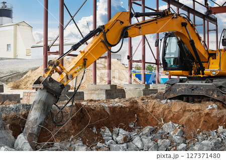 Construction site activity with excavator breaking concrete in broad daylight near industrial buildings Construction site activity with excavator breaking concrete in broad daylight near industrial buildings 127371480
