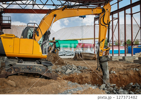 Excavator working on construction site during daytime in urban area with rocky terrain and industrial backdrop Excavator working on construction site during daytime in urban area with rocky terrain and industrial backdrop 127371482