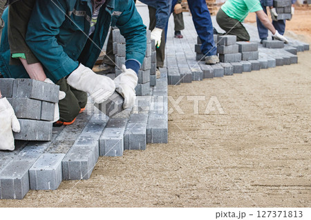 Workers laying grey paving stones during construction at a site in the afternoon light Workers laying grey paving stones during construction at a site in the afternoon light 127371813
