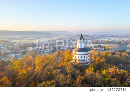 The elegant Baroque hunting chateau Humprecht stands above Sobotka, surrounded by vibrant autumn foliage, illuminated by the soft light of a tranquil sunrise. 127372711