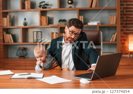 Busy father. Businessman in formal clothes is working indoors. With toddler 127374854