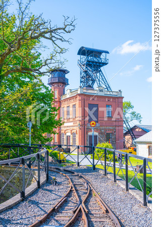 At Sevcinsky mine in Brezove hory, Pribram, Czechia, visitors explore a historical site with striking architecture and mining equipment surrounded by greenery and a clear blue sky. 127375556
