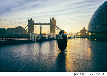 Morning light casts a warm glow over Tower Bridge, viewed from Queen's Walk. A modern sculpture reflects the sun as pedestrians enjoy the serene ambiance of London. Morning light casts a warm glow over Tower Bridge, viewed from Queen's Walk. A modern sculpture reflects the sun as pedestrians enjoy the serene ambiance of London. 127375561