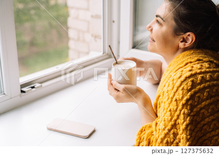 Woman smiling while holding coffee and looking out the window, enjoying tech-free time 127375632