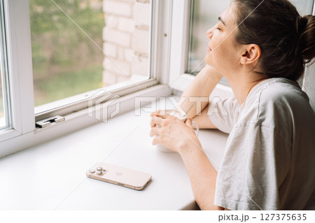 Smiling woman enjoying a hot drink by the window with a smartphone nearby 127375635