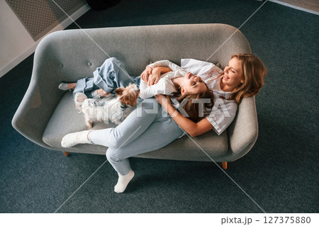Lying down on the sofa. Mother with her daughter and with cute dog is in domestic room Lying down on the sofa. Mother with her daughter and with cute dog is in domestic room 127375880