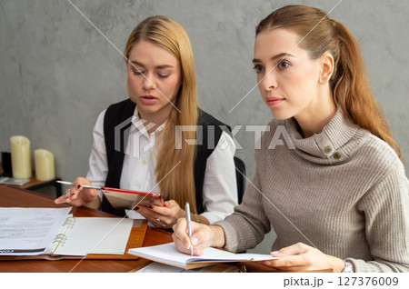 Two women engaged in a business meeting, taking notes and discussing strategies at a modern workspace. Two women engaged in a business meeting, taking notes and discussing strategies at a modern workspace. 127376009
