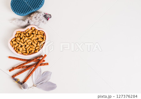 Bowl of cat food, treat, brush and toys on light background top view copy space 127376284