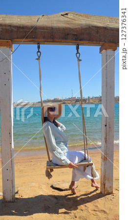 A female traveler in a straw hat rides on a swing against the backdrop of the sea, looking at the camera. A female traveler in a straw hat rides on a swing against the backdrop of the sea, looking at the camera. 127376341