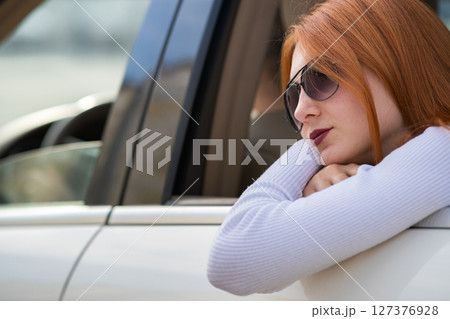 Young woman with red hair and sun glasses travelling by car. Passenger looking out of the rear window of a taxi in a city. 127376928