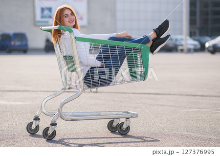 Young teenage happy girl sitting in supermarket shopping cart outdoors. 127376995