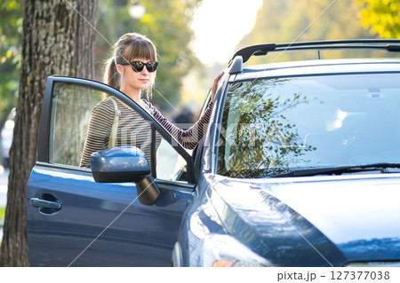 Young happy female driver standing near her car on a city street in summer. Travel destinations and transportation concept. Young happy female driver standing near her car on a city street in summer. Travel destinations and transportation concept. 127377038