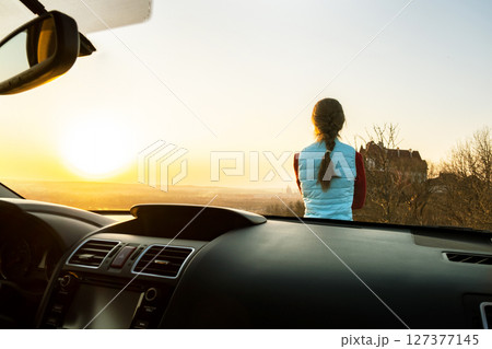 View from inside of a young woman standing near her car enjoying warm sunset view. Girl traveler leaning on vehicle hood looking at evening horizon. 127377145