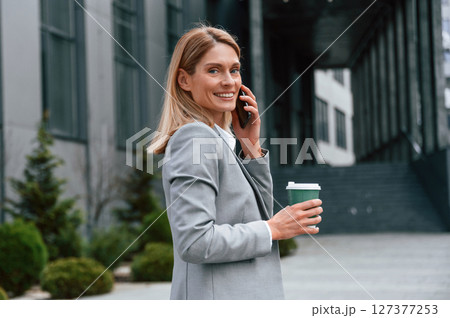Walking, holding cup of drink and talking by phone. Woman in formal clothes is outdoors near the business building Walking, holding cup of drink and talking by phone. Woman in formal clothes is outdoors near the business building 127377253