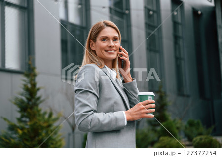 Walking, holding cup of drink and talking by phone. Woman in formal clothes is outdoors near the business building 127377254