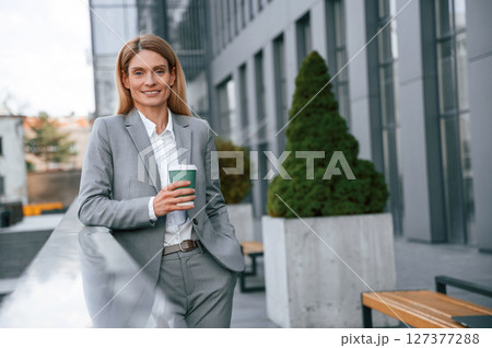 Smiling, holding cup of drink. Woman in formal clothes is outdoors near the business building 127377288