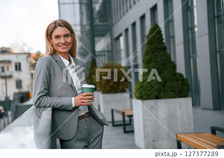 Smiling, holding cup of drink. Woman in formal clothes is outdoors near the business building 127377289