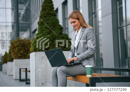 Sitting on the bench and using laptop. Woman in formal clothes is outdoors near the business building 127377290